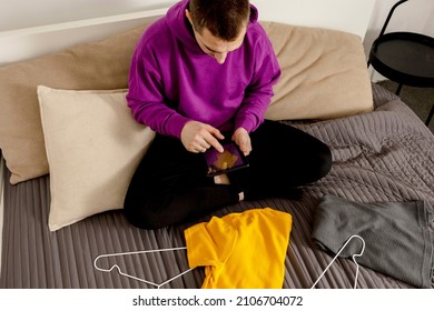 Man With Violet Hoodie Using Digital Tablet, Takes A Photo Of His Old Clothes To Sell Them Online. Selling On Website, E-commerce. Reuse, Second-hand Concept. Conscious Consumer, Sustainable Lifestyle