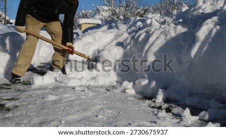 A man, visible without a face, clears large snowdrifts in front of a house with a wooden shovel in sunny weather. Manual snow removal from a house's surrounding area after a storm.