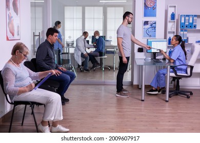 Man visiting hospital clinic for medical checking, giving radiography to assistant for new appointment at specialist doctor. Patients sitting in crowded waiting room filling form for consultation