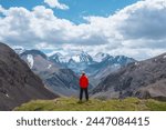 Man in vivid red jacket admire alpine scenery on sunlit green grassy hill near precipice edge. Guy on mountain pass enjoying view to few big snowy pointy peaks. Three large snow peaked tops far away.