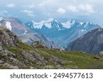 Man in vivid red jacket admire alpine scenery on green grassy hill among rocks near precipice edge. Guy with backpack on pass enjoying few big snowy pointy peaks. Three large snow peaked tops far away