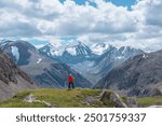 Man in vivid red jacket admire alpine scenery on sunlit green grassy hill near precipice edge. Guy on mountain pass enjoying view to few big snowy pointy peaks. Three large snow peaked tops far away.