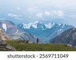 Man in vivid red jacket admire alpine scenery on green grassy hill among rocks near precipice edge. Guy with backpack on pass enjoying few big snowy pointy peaks. Three large snow peaked tops far away