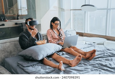 Man with VR headset and woman on laptop, modern couple multitasking in a high-rise apartment - Powered by Shutterstock