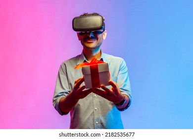 Man In VR Virtual Reality Glasses Holding Wrapped Gift Box And Smiling Joyfully, Giving Present To Camera, Celebrating Holiday. Indoor Studio Shot Isolated On Colorful Neon Light Background.