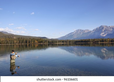 Man waist-deep in water fishing with reflections and mountains in background.