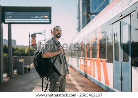 A man is waiting for a train on the platform. A young guy, a passenger with a backpack is standing on the platform waiting for the train