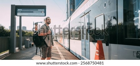 A man is waiting for a train on the platform. A young guy, a passenger with a backpack is standing on the platform waiting for the train