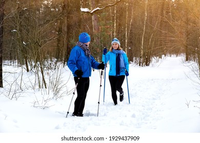 Man Waiting Woman On Walking With Nordic Walking Poles In Snowy Winter Park. Elderly Wife And Husband Doing Healthy Exercise Outdoors. Active Lifestyle After Retirement Concept.