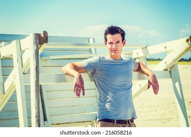 Man Waiting For You. Wearing A Gray T Shirt, Arms Resting On A Wooden Stick, A Young Handsome Guy Is Standing By A Wooden Structure On The Beach, Narrowing Eyes, Charmingly Looking At You. 