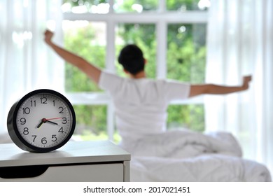 Man waking up in the morning sitting on bed and stretching, focus on alarm clock