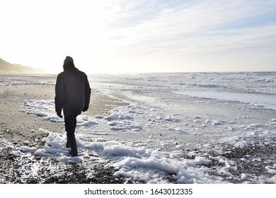 a man walking along the foamy beach against the sun light