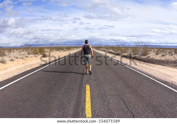 Man walking away from camera down center\
yellow line of black asphalt highway in barren Mojave Desert in\
California on sunny day with scattered\
clouds