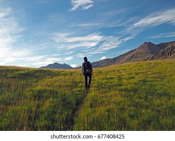 man walking with backpack through the meadow to the mountains with blue sky background