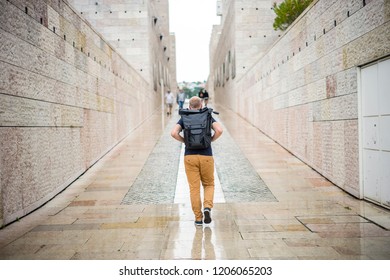 Man walking with backpack with tripod between high buildings during rainy weather