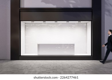 Man walking by evening empty shop window with podium inside for your product presentation on glowing white background in modern building area outdoors, mock up