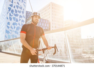 Man Walking And Carrying His Bike Over A Bridge. He Is Wearing An Helmet And Cycling Clothes. On The Background There Are Modern Glass Buildings. Sustainable Commuting Concept.