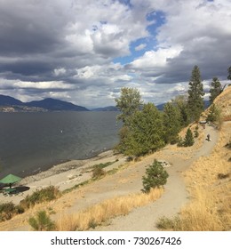 Man Walking Dog Uphill On Gravel Trail By Lake With Mountains Background.  Yellow Autumn Grass And Trees On Hill Overlooking Beach Along Lakeshore. Cloudy And Blue Sky Overhead.