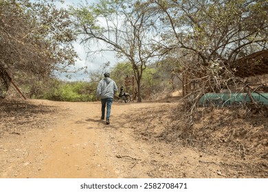 A man is walking down a dirt path in a forest. The path is narrow and surrounded by trees. The man is wearing a gray hoodie and jeans