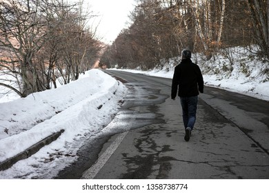 A man walking down an empty Winter road, lined by snow.
