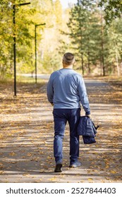 A man is walking down a path in the woods, wearing a blue sweater and blue pants. He is carrying a blue bag