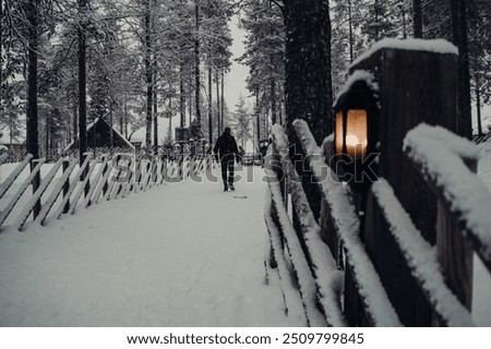Man walking down snowy path next to wooden fence and lanterns in Santa Claus' Village, Rovaniemi, Lapland, Finland