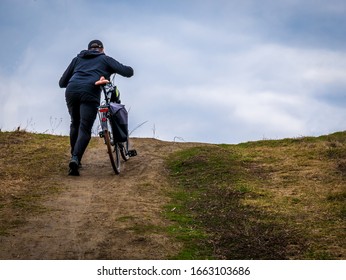 A man is walking up the hill with his bicycle. Man and his bicycle  on a cloudy day