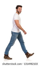 Man Is Walking In Jeans And White T-shirt, Looking At Camera And Smiling. Side View. Full Length Studio Shot Isolated On White.