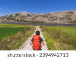 Man walking in nature path with backpack on shoulders exploring new places with amazing sight. He walks through fields along the banks of a river admiring the beauty of nature: meadow and mountains