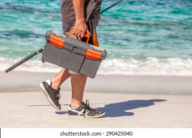 Man walking on the beach holding in one hand a tackle box and a fishing rod in the other.