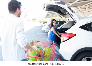 Man Walking With Shopping Cart Towards Girlfriend Standing By Car Trunk