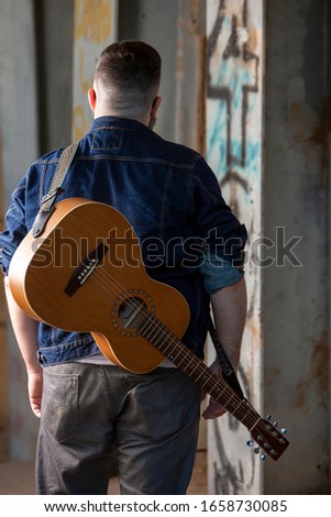 Man walking under the bridge with graffiti with an acoustic guitar strapped to his back wearing a blue denim shirt in Queen Creek Arizona