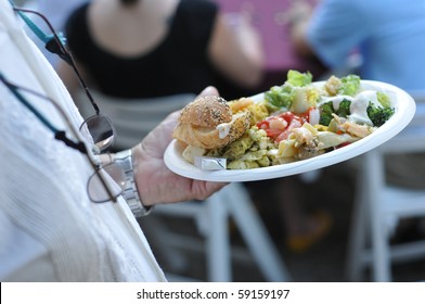 A Man Walks Back To His Table With His Paper Plate Filled With Food At A Party.