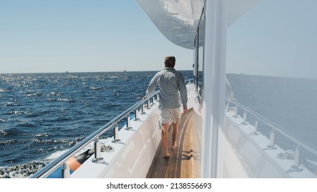 Man Walks On Moving Yacht Deck On Sunny Summer Day. Caucasian Man In Light Clothes, Barefoot On White Yacht Sailing By Blue Sea, Clear Sky Above Horizon. Luxury Lifestyle Vacation Private Sea Cruise