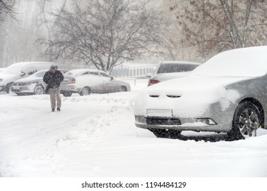 A man walks through the city during a snowstorm near snow-covered cars.