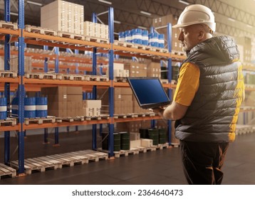 Man in warehouse. Logistics center employee. Worker with laptop in warehouse. Man inspects warehouse racks. Barrels and boxes are stored on pallets. Businessman with laptop in storage building