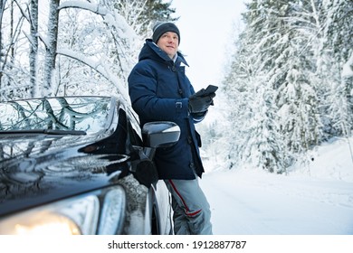 Man In Warm Winter Clothes Standing At The Car, Using Phone. Snowy Winter Country Road, Car Covered With Ice, Beautiful Forest Under The Snow. 