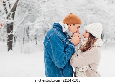 Man Warming Hands Of Woman With His Breath In Snowy Cold Winter Park. Loving Couple Walking Outdoors During Frosty Weather