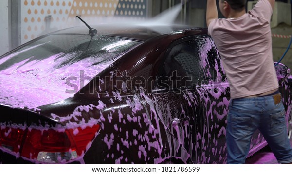 The man washes the car. Washes away pink foam with\
high pressure water