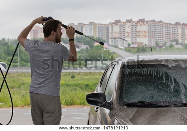 A man washes a car at a car wash,\
watering a car with a hose from a tap. City service for washing\
passenger cars. A young man with a beard shampooing a\
car