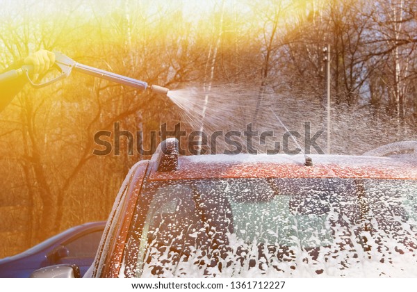 Man washes his orange car at car wash on clear\
sunny day. Cleaning with soap suds at self-service car wash. Soft\
yellow sunlight.