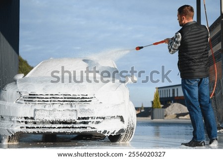 A man washes off the foam while washing the car. A vehicle is engulfed in soapy foam during a car wash, covering the tires, wheels, hood, and entire exterior of the car. Self-service car wash