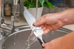 A Man Washes A Sharp Steel Dirty Kitchen Knife With A Sponge With Detergent In The Kitchen Sink Under Running Water. Gentle Hand Washing Of The Kitchen Knife. Cleaning Of Dirty Sharp Metal Products.