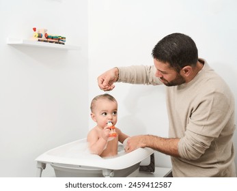 A man is washing a baby in a bathtub. The baby is holding a toy in his mouth - Powered by Shutterstock