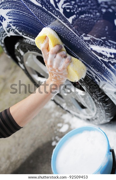 Man washing blue car with a yellow sponge and a\
bucket of soapy water.