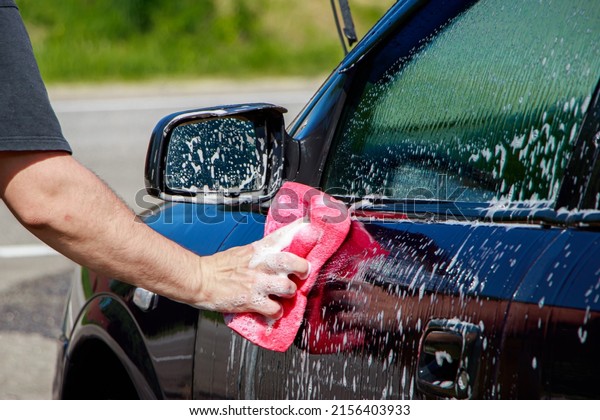 Man is\
washing car outside. Hand cleaning car in garage in front of house.\
Male hand holding pink sponge with\
foam.