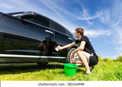 Man Washing Car with Soapy Sponge, Crouching Next to Green Bucket in Green Grassy Field on Bright Sunny Day with Blue Sky - Powered by Shutterstock