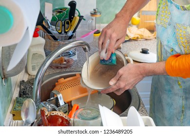 A Man Is Washing Dishes In Kitchen. He Is Using A Sponge And Dishwashing Product.  Hot Water Flows From The Tap