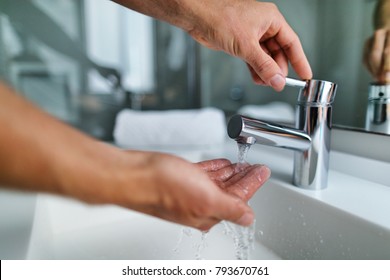 Man Washing Hands In Bathroom Sink At Home Checking Temperature Touching Running Water With Hand. Closeup On Fingers Under Hot Water Out Of A Faucet Of A Sink.