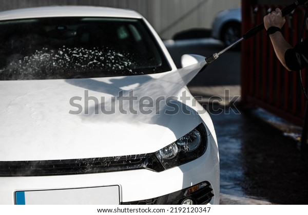 Man washing white car at contactless self-service\
car wash. Washing sedan car with foam and high-pressure water.\
Personal car care, cleaning outside. Nozzle spraying a jet of\
water, process of washing